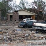 a person standing next to a several heavily damaged cars and a damaged house